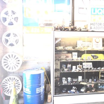 DTM Auto Salon - A glass display case showcases miniature car models and automotive accessories, positioned inside an auto parts shop. Behind the case, the storefront window features signs for LIQUI MOLY motor oils and additives, with visible vehicles and signage outside. The scene includes a blue barrel and wheel rims mounted on the wall, indicating an automotive retail environment. - Auto Repair Near Me - Pasadena, California