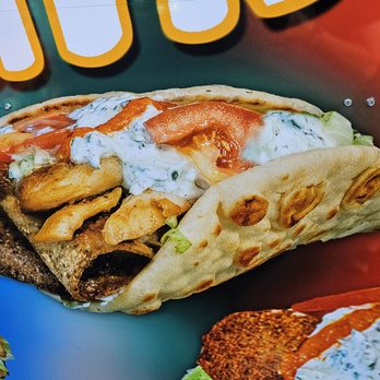 Crazy Gyro - A gyro sandwich with grilled meat, lettuce, tomato, and a creamy white sauce inside a toasted pita bread. The pita has brown spots from grilling. The sandwich is set against a colorful background with partially visible text, indicating a food or restaurant setting. - Mediterranean Near Me - Gresham, Oregon