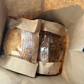 Small Axe Bread - The image shows two baked goods wrapped in paper, placed inside a brown paper bag. One appears to be a loaf of bread with a crusty surface, and the other looks like a darker pastry or bread with a glossy surface and some dark spots. The items are arranged side by side within the bag, which is open at the top, revealing the baked goods inside. - Bakeries Near Me