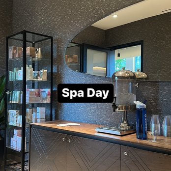 Equinox - Berkeley - The charcoal water and items available for purchase at the Spa entrance. - The image shows a spa reception area with a dark textured wall and a curved architectural feature. A glass display shelf on the left holds various skincare and wellness products. The wooden counter in the foreground has a water dispenser, a few glasses, and some papers. A window behind the counter provides natural light and a view outside. The space appears modern and well-organized. - Gyms Near Me - Berkeley, California