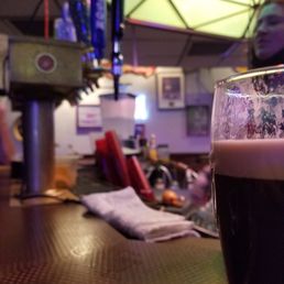 A dark beer in a pint glass is placed on a bar counter, with a blurred background showing bar equipment, a person, and colorful lighting.