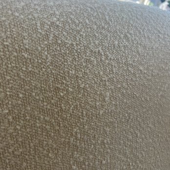Beeclean - This is what it looks like before. - The image shows a close-up of a beige fabric surface with a textured, granular appearance, likely upholstery or carpet material. The background includes a window with natural light coming through, and some objects or decorations are visible outside the window, slightly blurred. The focus is on the fabric's detailed surface texture. - Carpet Cleaning Near Me