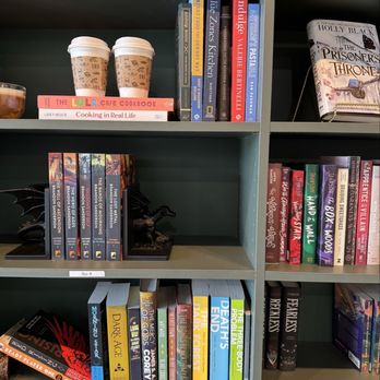 Book Bar - Multiple bookshelves contain a variety of books, including fiction, non-fiction, and cookbooks, with some labeled as 'Sci-fi.' Two coffee cups are placed on the top shelf, along with a small dragon figurine on the middle shelf. The setting appears to be a bookstore or library with a cozy atmosphere, featuring a curated selection of books and decorative items. - Sandwiches Near Me - Mokena, Illinois