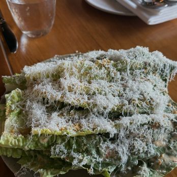 PASTA - Romaine, peperoncino sott'olio, parmigiano reggiano, pangrattato - A Caesar salad with lettuce and grated cheese is served on a ceramic plate on a wooden table, with a glass of water and tableware nearby. - Italian Near Me - Miami, Florida