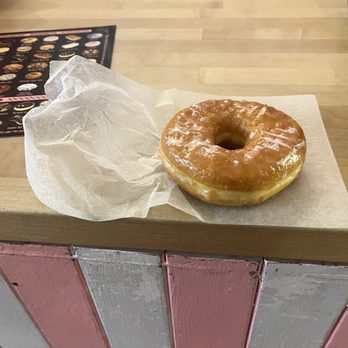 Voodoo Doughnut - The whole glazed yeast. - A single glazed donut with a shiny, smooth icing surface rests on a piece of parchment paper placed on a wooden counter. In the background, a menu with various donut options is partially visible, indicating a donut shop setting. - Donuts Near Me - Austin, Texas