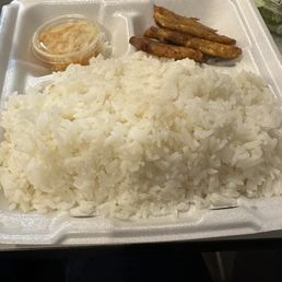 Pikliz - A foam takeout container holds a serving of white rice, fried plantains, and a small cup of sauce, representing Haitian cuisine. The rice is piled on one side, with fried plantains placed on the upper right corner. The small cup of sauce is positioned near the rice, and a portion of leafy greens is visible in the background. The container is open, showing the food inside.