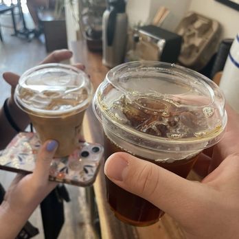 Café Domestique - Latte and cold brew - Two people are holding cups of iced coffee with plastic lids and straws inside a café. One person has a floral phone case and painted nails, while the other has a plain hand. The background shows a wooden counter with a thermos, a small black appliance, and a paper bag, with a blurred view of the café interior and seating area. - Coffee & Tea Near Me - Madison, Wisconsin
