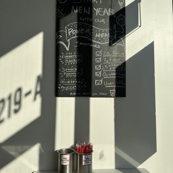 Smoothie King - a menu on the wall - A black chalkboard menu lists smoothie flavors and nutritional information, including protein, fiber, vitamins, and sugar content, with sunlight casting shadows on the wall. Two stainless steel containers filled with red straws are placed on a white table below the menu, with a small black tray and a round metallic object nearby. - Health Markets Near Me - Frederick, Maryland