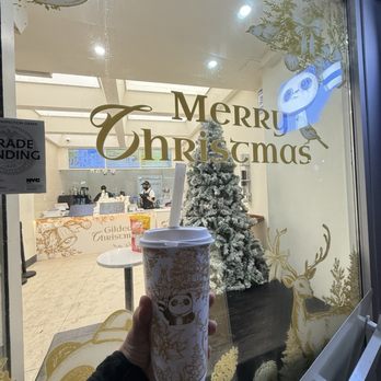 Tea By Do - A hand holds a paper cup with a straw in front of a decorated window featuring Christmas and panda-themed decorations, with a Christmas tree inside. - Bubble Tea Near Me - Flushing, New York