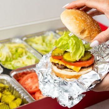 Five Guys - A close-up photo of a person putting the top bun of their cheeseburger in front of a Five Guys catering box of toppings. - Burgers Near Me - Fort Smith, Arkansas Five Guys - A close-up photo of a person putting the top bun of their cheeseburger in front of a Five Guys catering box of toppings. - Burgers Near Me - Fort Smith, Arkansas