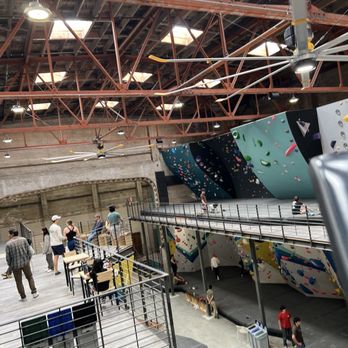 The Oaks - The image shows an indoor rock climbing gym with high ceilings and exposed structural beams. Several climbing walls with various angles and colorful holds are visible, along with a padded floor area. People are seen standing, sitting, or preparing to climb, with some on the upper platform and others near the climbing walls. The space includes safety equipment and seating areas, indicating a facility designed for indoor climbing activities. - Rock Climbing Near Me - Berkeley, California