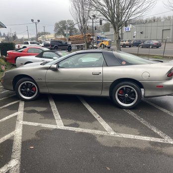 Summers Car Company Classics - A silver Chevrolet Camaro SS with a black roof is parked in a parking lot, occupying two parking spaces. The car features black wheels with red brake calipers and a sleek, sporty design. Surrounding the Camaro are other vehicles, including a red sports car and a white car, with a business building and trees in the background. The parking lot has white painted lines and some patches of water on the asphalt. - Car Dealers Near Me - Eugene, Oregon