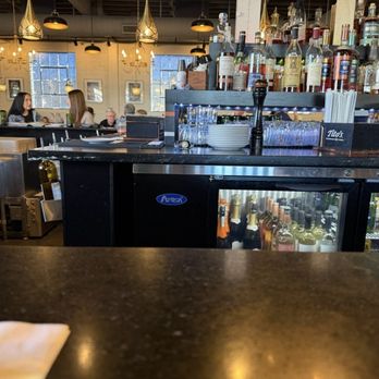 M’tucci’s Bar Roma - Bar - Interior view of a bar in a restaurant, featuring liquor bottles on shelves, glasses, and a refrigerator with beverages, with patrons in the background. - Cocktail Bars Near Me - Albuquerque, New Mexico