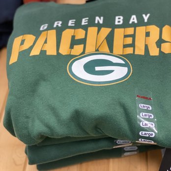 Scheels - A green sweatshirt with the Green Bay Packers logo and text is folded on a wooden surface. The sweatshirt displays the team's name, logo, and multiple size labels indicating 'Large.' The background includes other clothing items and a wooden table, suggesting a retail or shopping environment. - Hunting & Fishing Supplies Near Me - West Des Moines, Iowa