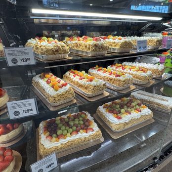H-E-B - A glass display case at H-E-B in Lakeway, TX, showcases an assortment of cakes, including fruit-topped sheet cakes and strawberry cakes, with price tags visible. - Drugstores Near Me - Lakeway, Texas