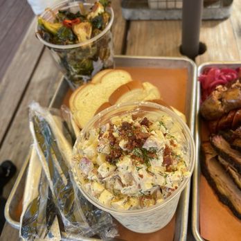 Truth BBQ - Potato salad & Brussel Sprouts with potato bread!! - A cup of potato salad with bacon, herbs, and vegetables is placed on a tray alongside sliced bread and a cup of roasted vegetables. - Barbeque Near Me - Brenham, Texas