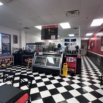 Sonsons Pasty - The photo depicts the interior of Sonsons Pasty Company, a bakery with a black and white checkered floor, black chairs with red cushions, and a red accent wall decorated with framed pictures. The counter area includes display cases, a menu hanging from the ceiling, and a yellow caution sign on the floor. The space appears to be a casual dining area with self-service options and a small seating arrangement. - British Near Me - Mesa, Arizona