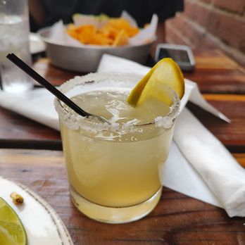 Bakersfield Mass Ave - Skinny margarita - A salted rim margarita with a lemon wedge is on a wooden table, with a bowl of chips, a glass of water, and a phone in the background. - Bars Near Me - Indianapolis, Indiana