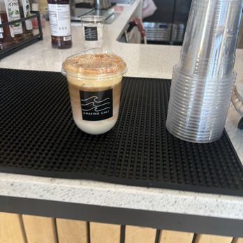 Chasing Cali Collective - A layered coffee drink in a clear plastic cup features a white milk layer at the bottom, a dark coffee layer in the middle, and a frothy top. The cup has a black label with a white logo reading 'Chasing Cali.' The drink is placed on a black rubber mat on a white countertop, with a stack of clear plastic cups to the right. The background shows a cafe setting with various bottles, containers, and equipment. - Coffee & Tea Near Me - Cincinnati, Ohio