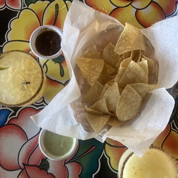 Armadillo - A basket of tortilla chips with three dipping sauces—black, green, and yellow—on a vibrant floral tablecloth, accompanied by two margaritas. - Mexican Near Me - Kingston, New York