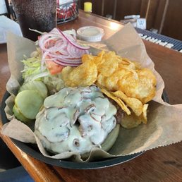 Mushroom & Swiss - A plate features a burger topped with a creamy mushroom and Swiss cheese sauce, accompanied by sliced onions, pickles, and fried onion rings. A glass of soda with ice is visible in the background. The meal is served on a paper-lined tray at a restaurant or pub setting, with condiments and a wooden counter visible.