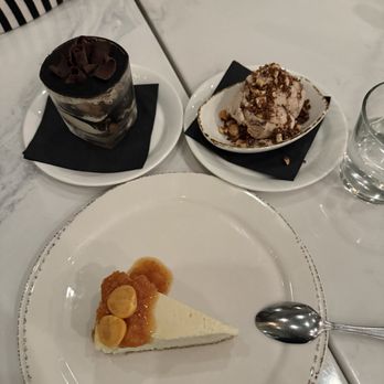 Marina - Tiramisu, hazelnut gelato, Blue Cheese Cheesecake - The image shows a marble table with three desserts and a glass of water. A slice of cheesecake with fruit topping is on a white plate, accompanied by a spoon. Two small dishes contain a chocolate dessert with chocolate shavings and a scoop of ice cream with nuts. A person's hand and part of their face are visible in the background, along with a folded napkin. - Italian Near Me - Pasadena, California