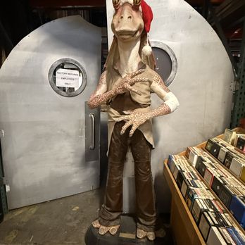 Factory Records - A figure with a dinosaur head and humanoid body, dressed in vintage-style clothing, stands in front of a metal door labeled 'Factory Records Employees Only' at a record store. - Vinyl Records Near Me - Dover, New Jersey