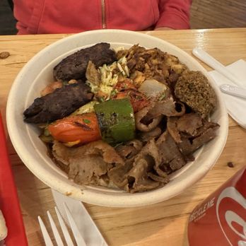 Rumi Middle Eastern Grill - *NEW* Mixed Shawarma Plate - A disposable bowl holds a variety of Middle Eastern foods, including grilled meats, roasted vegetables, falafel, and rice, with some salad and sauces. The meal is placed on a wooden table, with a plastic fork, knife, and napkin nearby. A person wearing a red hoodie is seated behind the table, and a Coca-Cola can is partially visible in the corner of the image. - Mediterranean Near Me - Chicago, Illinois