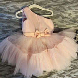 A pink dress with a sleeveless design and pleated bodice is hanging on a white hanger. The dress features multiple layers of tulle fabric creating a full skirt, with a satin bow at the waist. It is displayed against a textured, patterned background, likely on a bed or surface. The dress appears suitable for a young girl, possibly for a special occasion or dress-up.