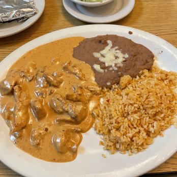Playa Chac Mool - Pollo Chac - Mool - A plate of Mexican cuisine includes seasoned rice, beef stew topped with cheese, and chicken in a creamy sauce. In the background, there are small bowls of guacamole and salsa, along with foil-wrapped items on a wooden table. The setting suggests a casual dining environment with traditional Mexican dishes. - Mexican Near Me - St. Augustine, Florida