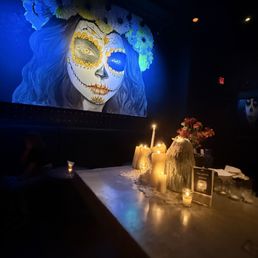 Bar area - A mural of a woman with sugar skull makeup, including painted facial features and floral designs, is painted on a dark wall. The mural shows her with long, wavy hair and a large floral headdress. Below the mural, a table is decorated with lit candles, a large stone-like object, and a bouquet of pink flowers, creating a dimly lit ambiance.