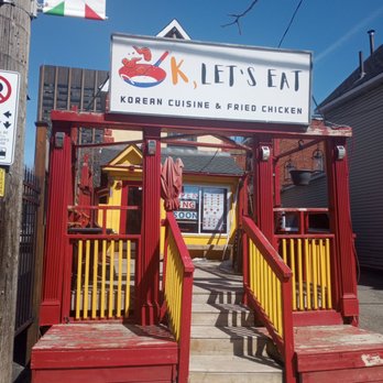 K, Let's Eat - Front of the restaurant - The image shows the storefront of K, Let's Eat, a restaurant specializing in Korean cuisine and fried chicken. The entrance is accessed via a small staircase with red and yellow railings. A signboard above the entrance displays the restaurant's name and a graphic of fried chicken, along with the description 'Korean Cuisine & Fried Chicken.' The building behind the staircase is painted yellow, and there is a window with a menu and an 'Open' sign. The surrounding area includes a brick sidewalk and neighboring buildings, with a clear blue sky overhead. - Korean Near Me - Ottawa, Ontario
