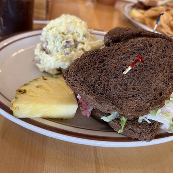 Christy Mae's Restaurant - Reuben - really great - A plate features a sandwich made with dark rye bread, filled with lettuce and sliced meat, with toothpicks securing the sandwich. Alongside, there is a scoop of potato salad with visible bits of bacon or ham and a slice of pineapple. The background shows a glass of dark soda and another plate of food, indicating a casual dining setting. - Sandwiches Near Me - Albuquerque, New Mexico