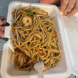 Lo Mein - A foam takeout container holds stir-fried lo mein noodles with vegetables, shrimp, and chicken, served on a gray table.