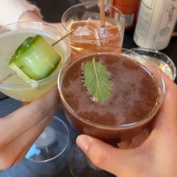 a close up of two people holding cocktails - Two people are holding three cocktails in front of a bar counter. The drinks include a green cocktail with a cucumber garnish, a brown cocktail with a leaf garnish, and an orange-colored drink with a straw. The background shows various bottles and bar equipment, indicating a bar setting.