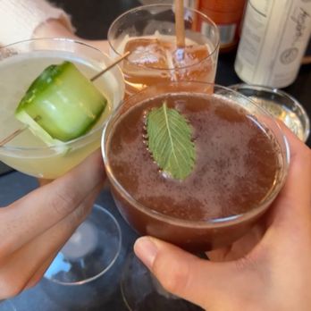 Nook - a close up of two people holding cocktails - Two people are holding three cocktails in front of a bar counter. The drinks include a green cocktail with a cucumber garnish, a brown cocktail with a leaf garnish, and an orange-colored drink with a straw. The background shows various bottles and bar equipment, indicating a bar setting. - Cocktail Bars Near Me - Somerville, Massachusetts