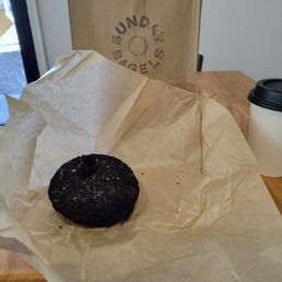 A black sesame harissa bagel, toasted with butter - A black sesame harissa bagel, toasted with butter, sits on parchment paper on a wooden table, with a coffee cup and paper bag in the background.