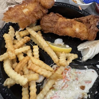Culver's - Fish and fries! - The image shows a meal with two pieces of fried fish fillets, one larger and one smaller, on a black tray. Accompanying the fish are crinkle-cut fries, a portion of coleslaw, a lemon wedge, and a small container of tartar sauce. There is also a cup of creamy dressing and some condiment packets visible on the table. The setting appears to be casual, likely at a fast-food or casual dining restaurant. - Fast Food Near Me - Mesa, Arizona
