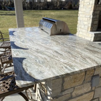 Stone Statements - An outdoor stone countertop with a polished, patterned surface is mounted on a stone base, with chairs nearby and a grill in the background. - Building Supplies Near Me - Cincinnati, Ohio