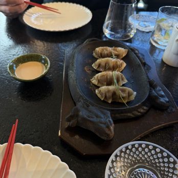 Makoto Vail - WAGYU DUMPLING - The image shows five dumplings placed on a fish-shaped hot plate, likely served as part of Japanese cuisine. The hot plate is on a dark table, with a small bowl of dipping sauce nearby. In the background, a person is holding chopsticks over an empty white plate, and there are glasses of water and a glass with a lemon slice. The setting appears to be a Japanese restaurant with a modern interior. - Sushi Bars Near Me - Vail, Colorado