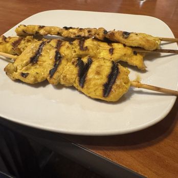 Thai Tip - A-7 chicken Satay 3pcs. - Three chicken satay skewers with grilled marks and a yellow marinade are placed on a white oval plate. The skewers are arranged parallel, with the sticks extending beyond the plate's edge. The chicken pieces appear cooked and seasoned, with some sauce drizzled on top. The plate is set on a wooden table, suggesting a restaurant setting, likely serving Thai cuisine. - Thai Near Me - Albuquerque, New Mexico
