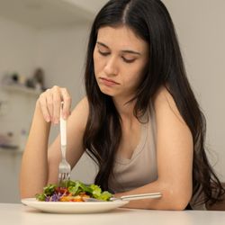 Connections Wellness Group - Young woman looking down at a plate of food with a serious or reluctant expression, symbolizing challenges with eating behavi...