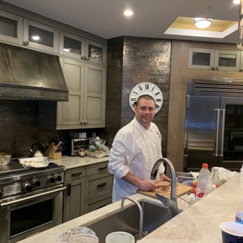 Vail Catering Concepts - Jonathan prep'ing for private dinner in Vail Village - A male chef wearing a white chef's coat is standing at a kitchen counter, preparing food with a knife. The kitchen features stainless steel appliances, including a large refrigerator, and cabinetry with a gray finish. The background includes a clock on a brick wall, and the counter has various items such as bottles, containers, and utensils. The space is well-lit with ceiling lights and a decorative light fixture. - Caterers Near Me - Minturn, Colorado