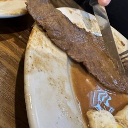A piece of cooked beef steak is placed on a white plate with sauce underneath. A person is cutting the steak with a knife, and a fork is held in the other hand. Part of a small bowl with some food and sauce is visible in the background. The setting appears to be a dining table with a wooden surface.