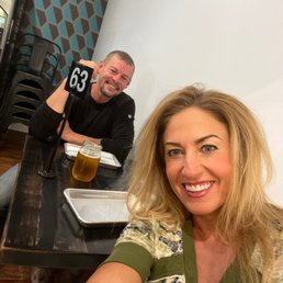 A woman with blonde hair is smiling and taking a selfie in a restaurant. Behind her, a man is sitting at a table, smiling at the camera, with a table number 63 sign and a jar of drink in front of him. The background includes a patterned wall and stacked chairs. The table has a tray and some napkins, indicating a casual dining setting.