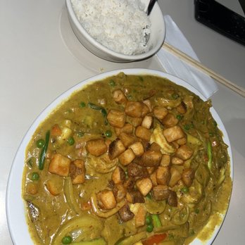 Pho Leo - X54. Curry (Spicy). Waiting Period 15-20 min. - A large white plate contains a curry dish with fried tofu cubes, green peas, and vegetables in a thick sauce. A separate bowl of white rice with a spoon is placed nearby. The meal is set on a white table with chopsticks, a napkin, a water bottle, and a smartphone visible in the background. - Vietnamese Near Me - Mesa, Arizona