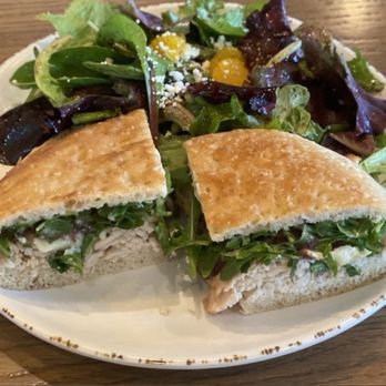 Urbane Cafe - A sandwich with toasted bread, filled with leafy greens and sliced chicken, is cut in half and placed on a white plate. Accompanying the sandwich is a mixed green salad with various lettuce leaves, yellow cherry tomatoes, and crumbled cheese. The plate is set on a wooden table, and the image captures the meal from a top-down perspective. - Salad Near Me - Menifee, California