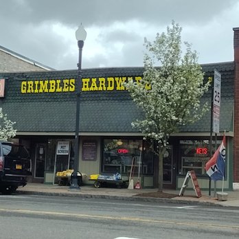 Grimbles Hardware - The image shows the storefront of Grimbles Hardware, with a large yellow sign displaying the store's name. In front of the store, there is a blooming tree with white flowers, and a street lamp is positioned nearby. The storefront has multiple signs, including one for pet screens, keys, and an illuminated 'OPEN' sign. The sky above is cloudy, indicating overcast weather. The sidewalk features some equipment and a flag, with a street and traffic lines visible in the foreground. - Keys & Locksmiths Near Me - Lockport, New York