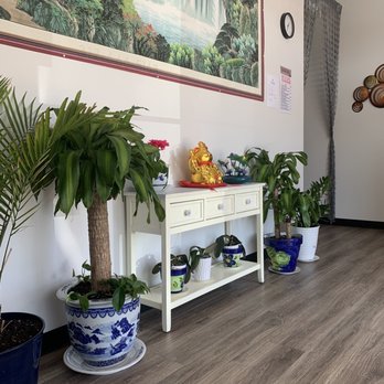 Luna Spa - Inside - The image shows an indoor space with a white console table against a white wall, decorated with potted plants of various sizes and styles. A large painting of a landscape with waterfalls and mountains hangs above the table. Traditional Chinese decorative items, including a gold figurine and small vases, are placed on the table. The floor is covered with wood-like flooring, and a clock and some papers are attached to the wall. Curtains are visible on the right side of the image. - Massage Near Me - Plainfield, Indiana