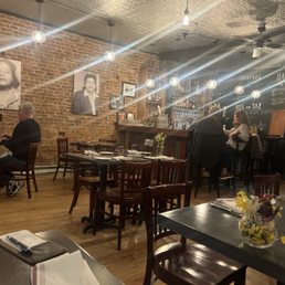 The image shows the interior of a restaurant with exposed brick walls and warm lighting from hanging pendant lights. There are wooden tables and chairs, some set with napkins and utensils. A bar area with shelves of bottles and glasses is visible in the background, with patrons seated at the bar. Black-and-white photographs adorn the brick wall, and a chalkboard menu is partially visible. The space has a cozy, rustic atmosphere suitable for dining.